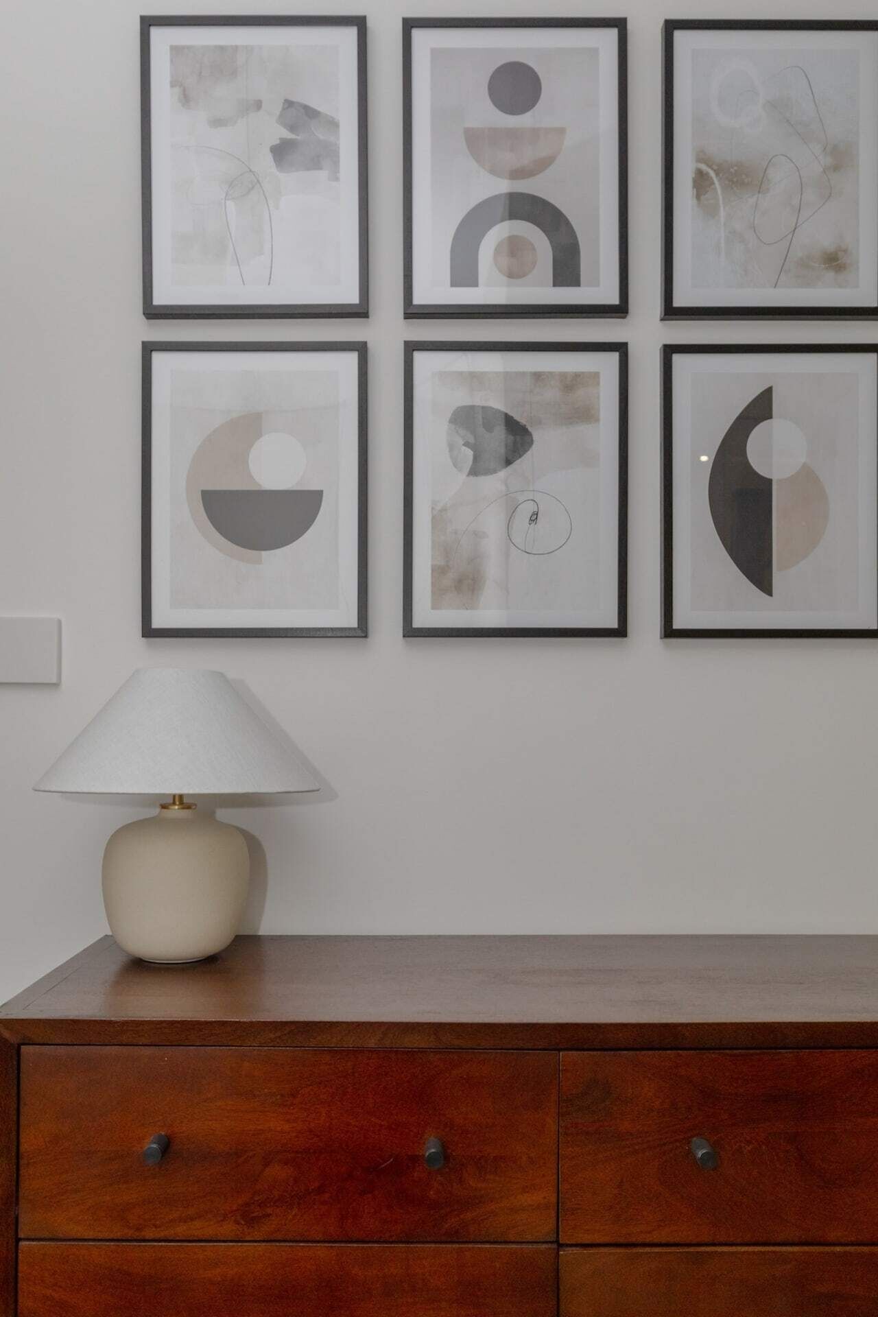 Un mur orné de six œuvres d’art abstraites encadrées, une lampe blanche et un meuble en bois doté de trois tiroirs.