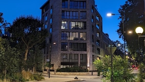 A multi-story building with a modern architectural design, featuring a mix of glass and stone facade, surrounded by greenery and street lighting. - Elegant & Serene 2BD Flat W/roof Terrace Hoxton! (London)