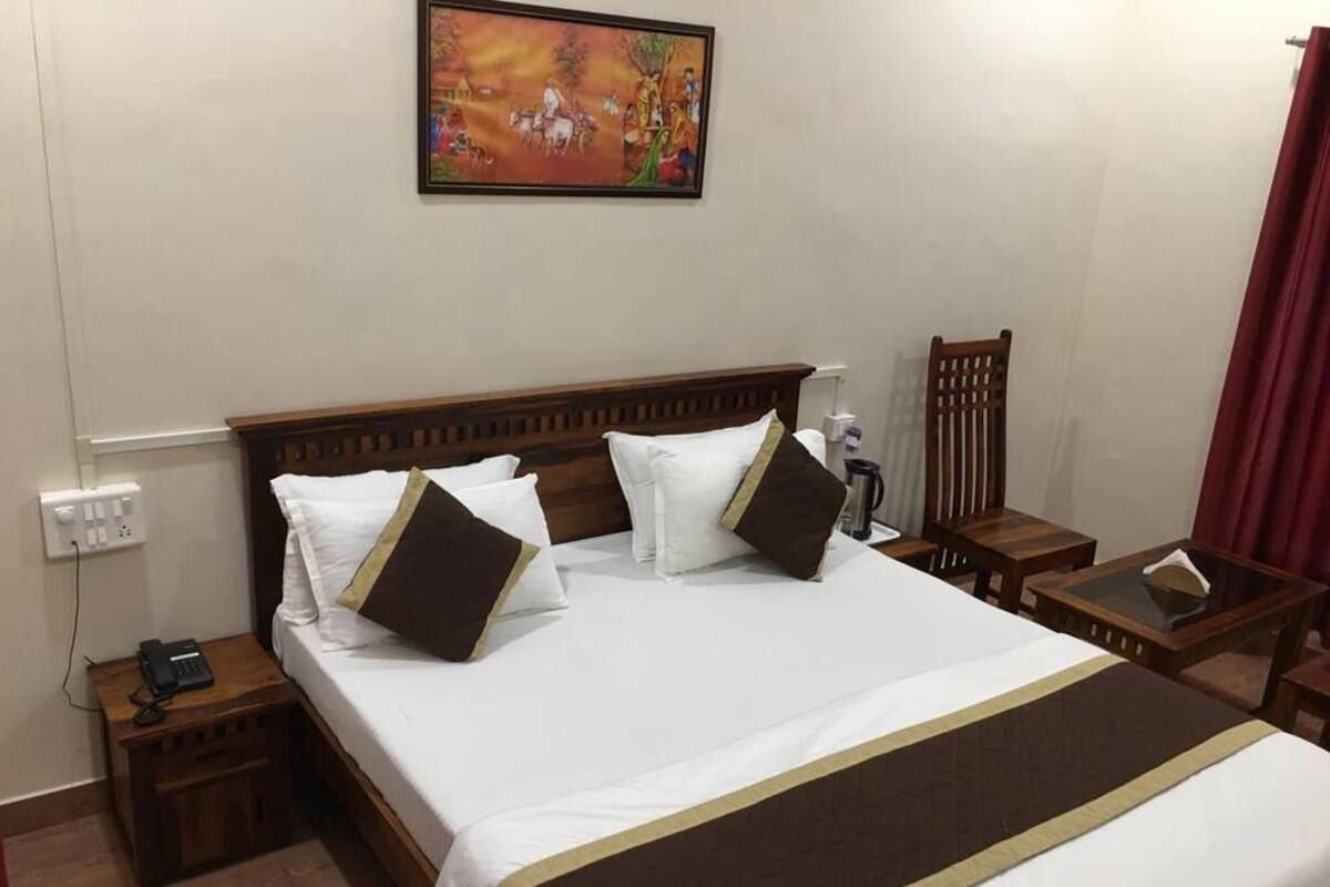 Una cama bien hecha con cabecera marrón, ropa de cama blanca y un cojín decorativo marrón. Hay una mesita de noche con una lámpara y un teléfono. Un cuadro cuelga de la pared sobre la cama.
