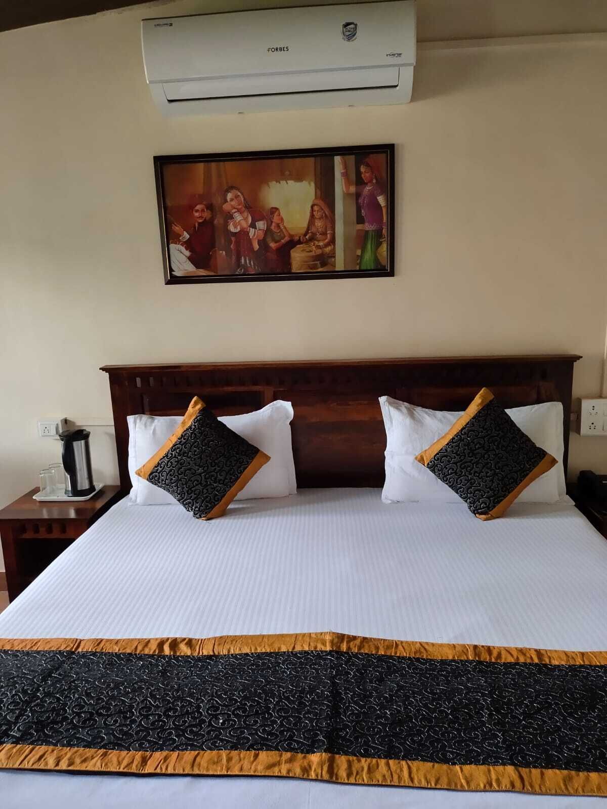 Habitación de hotel con una cama grande, dos almohadas con estampado negro y dorado, un cabecero de madera, un cuadro sobre la cama y un equipo de aire acondicionado en la pared.