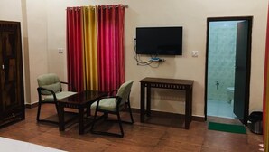 A room with a TV mounted on the wall, a small table, two chairs, and a window with curtains. - Rose Cottage A Heritage Retreat (Abu Road)