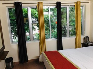 Une chambre d’hôtel avec un lit, une fenêtre avec des rideaux, une poubelle et une petite table avec une bouilloire.