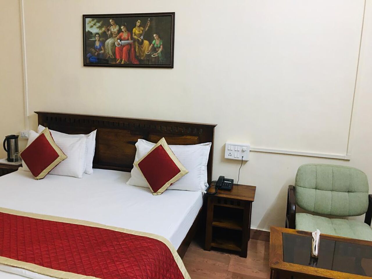 A hotel room with a large bed, two red pillows, a wooden headboard, a nightstand with a phone, a green armchair, and a painting on the wall.