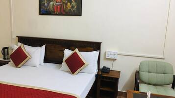 A hotel room with a large bed, two red pillows, a wooden headboard, a nightstand with a phone, a green armchair, and a painting on the wall.