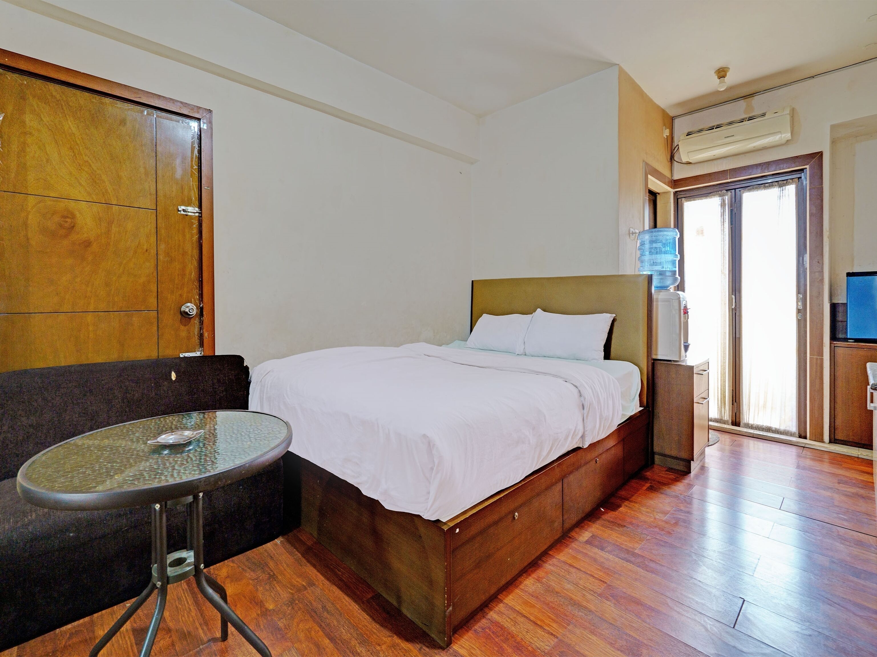 A hotel room with a wooden bed, a glass table, a sofa, and a water dispenser.