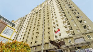 A high-rise building with multiple balconies, a flag, and some greenery in the foreground.