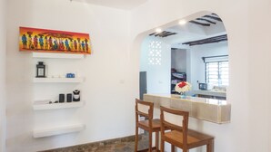 Un comedor con suelo de piedra, paredes blancas, un cuadro y una cocina visible a través de un arco.