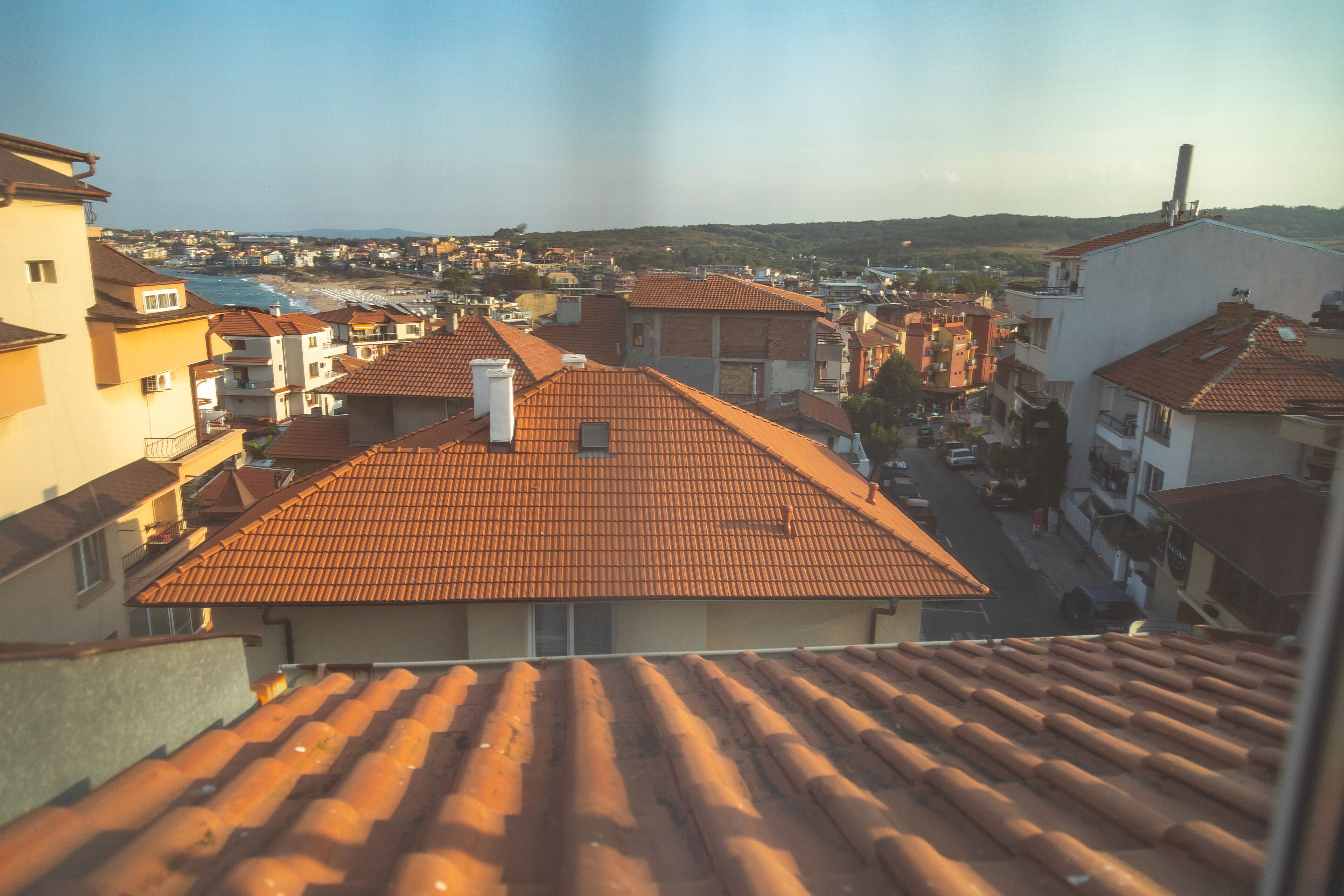 A residential area with terracotta-tiled roofs, a clear sky, and a distant view of a beach.