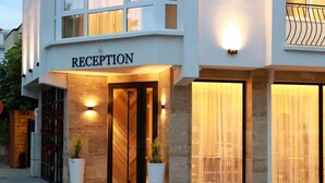 Zona de recepción de hotel, con un cartel que dice 'RECEPTION', ventanas amplias con cortinas y dos plantas en macetas.