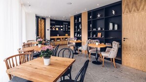 Un café moderno con mesas y sillas de madera, una estantería con objetos decorativos y una amplia puerta de madera.