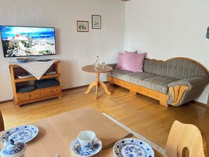 Ein Wohnzimmer mit einer Couch, einem kleinen runden Tisch, einem an der Wand montierten Fernseher und einer Holzkommode.