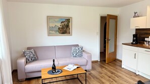 A living room with a sofa, a coffee table, a painting, and a kitchen area with cabinets and a coffee machine.