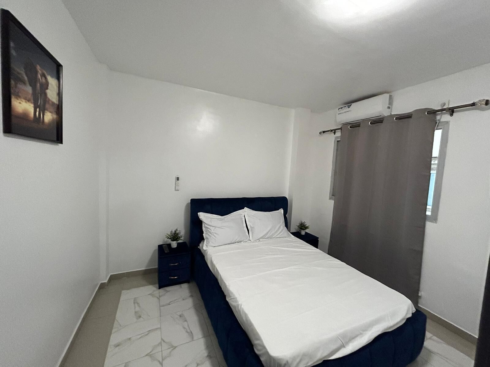 A bedroom with a bed, a nightstand, a small plant, and a window with curtains.