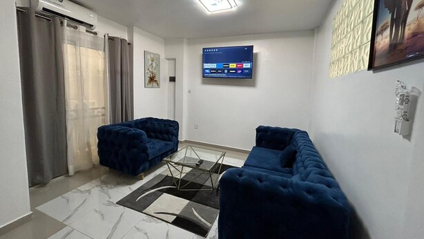 A modern living room with two blue sofas, a flat-screen TV, and a patterned rug. - Studio Meublés Haut Standing À Bonamoussadi (Douala)