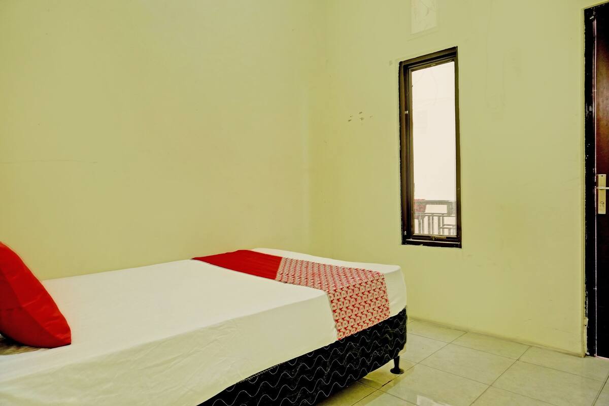 Ein Einzelbett mit weißer Matratze, roter, gemusterter Bettwäsche und einem schwarzen Bettgestell. Ein Fenster mit schwarzem Rahmen ist an der Wand erkennbar.