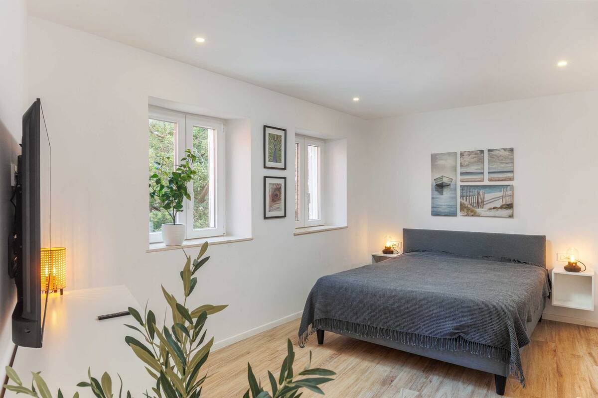 A bedroom with a bed, bedside tables, a TV, windows, and wall art.