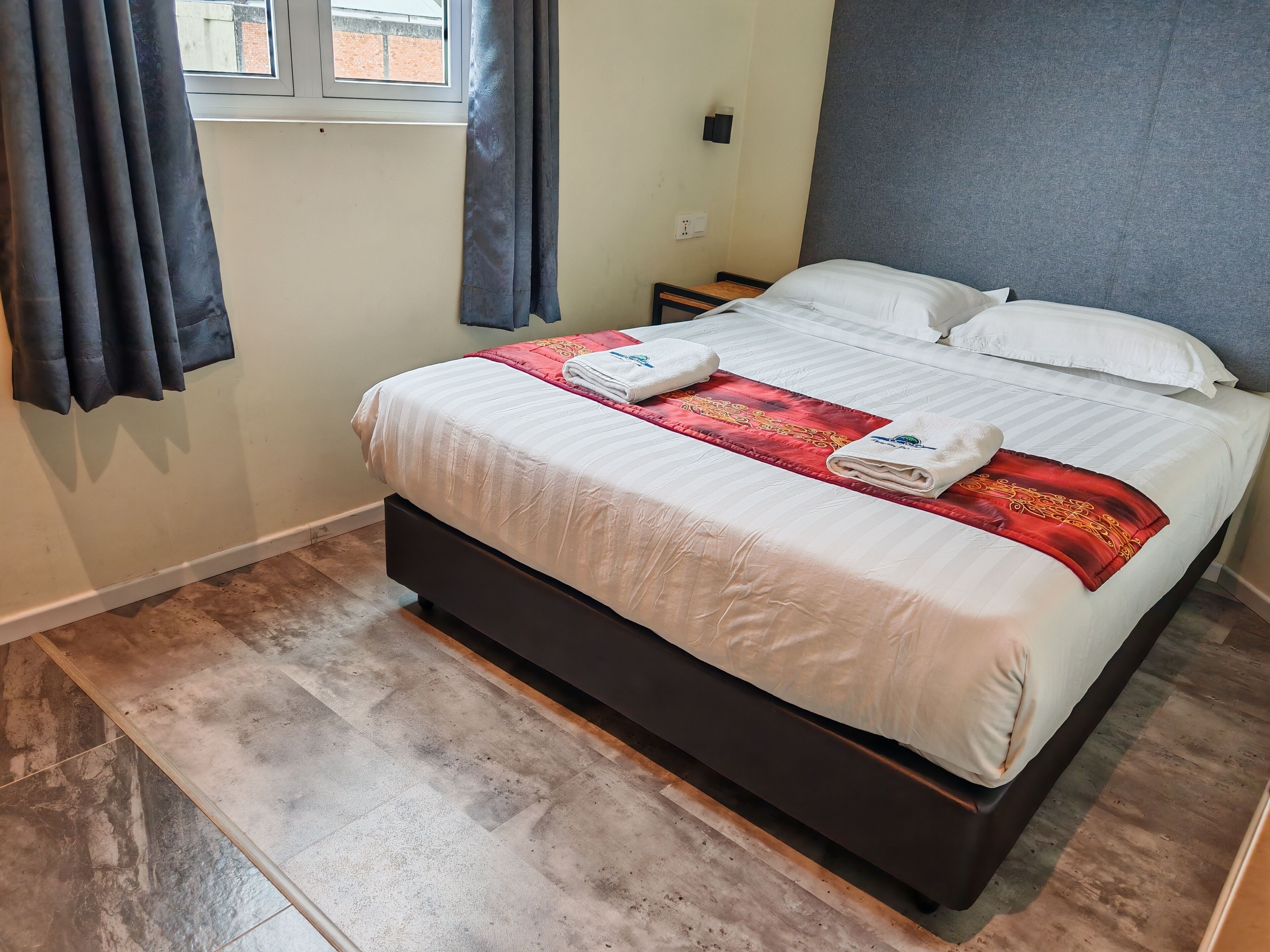 Een keurig opgemaakt bed met een wit-rode dekbedovertrek, twee handdoeken en een klein nachtkastje met een lamp.