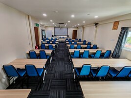 A conference room with rows of blue chairs and wooden tables, a projector screen, and a fire extinguisher.