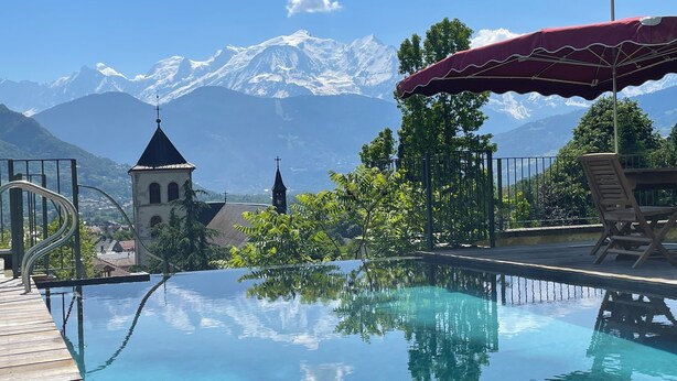 A clear pool with a wooden deck, a red umbrella, and a mountain view.