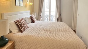 A bedroom with a large bed, a ceiling fan, a nightstand, a lamp, and a window with curtains.