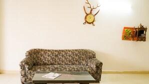A living room with a patterned sofa, a wooden coffee table, a wall-mounted clock, and a small shelf with decorative items. - Roomshala 002 Rose Residency (New Delhi)
