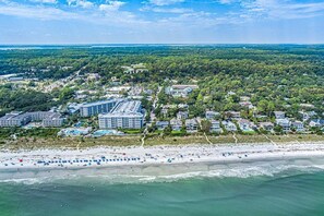An aerial view of a coastal town with a beachfront, including a hotel with multiple buildings and a beach with umbrellas. - Newly Renovated • 60 StepstoBeach • Pool • FirePit * Screen Patio * Walk to Coligny • (Hilton Head Island)