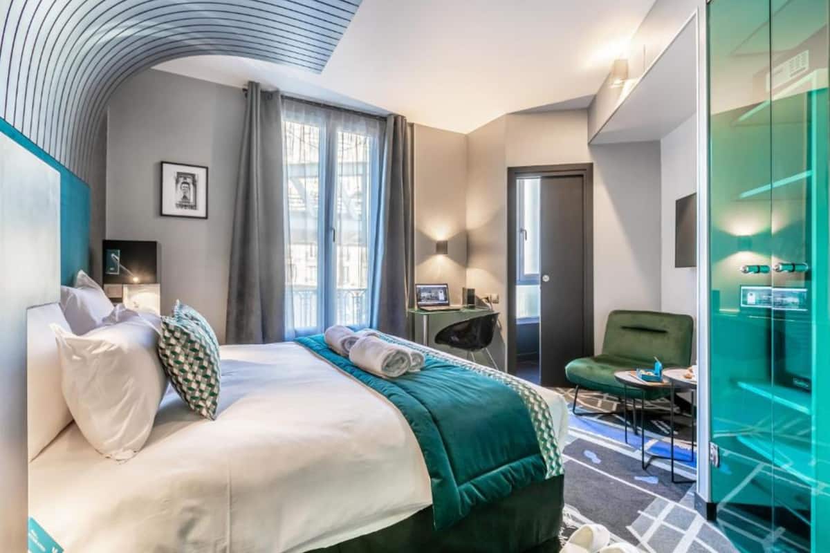 A modern hotel room with a curved ceiling, a large bed with white and green bedding, a desk with a chair, and a glass partitioned area.
