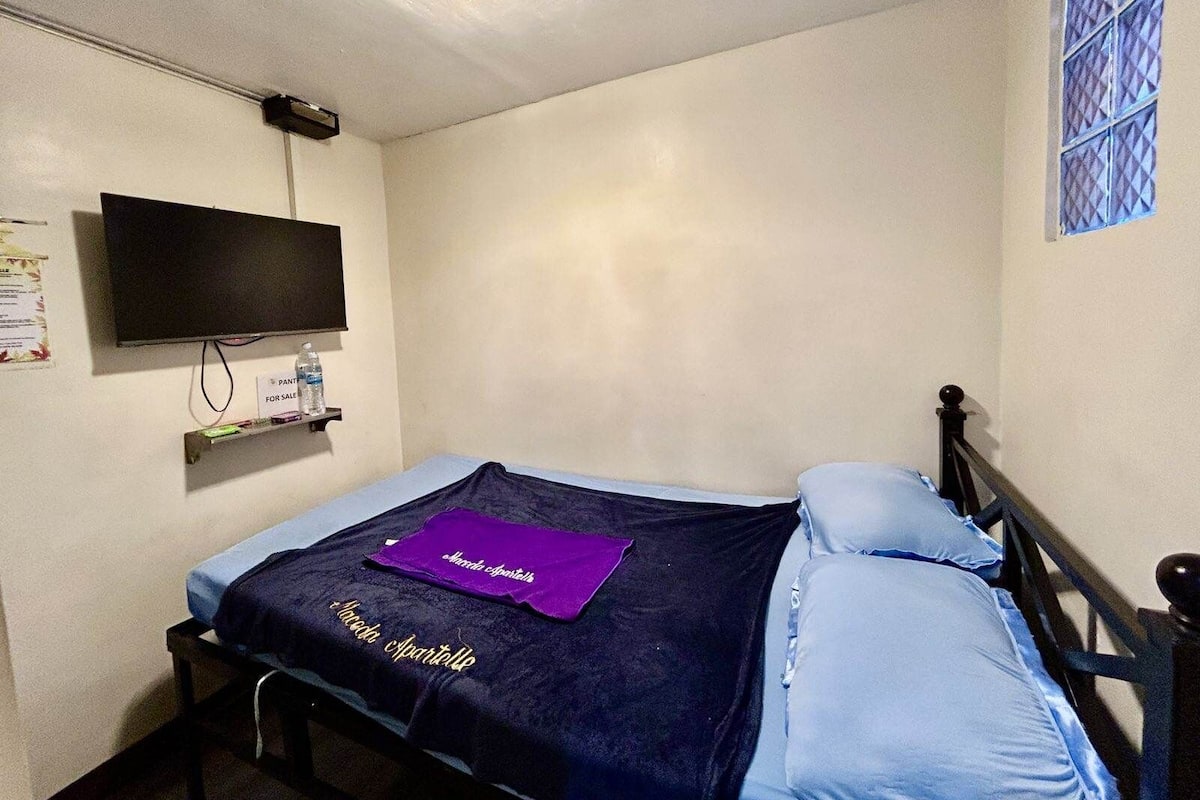 A small, simple room with a bed, a television mounted on the wall, and a window with a patterned blind.
