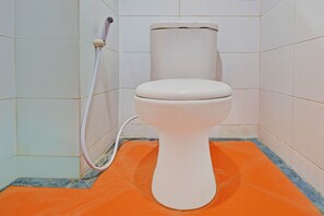 Un inodoro con tanque y asiento blancos, una manguera rosa y un tapete naranja en un baño con paredes de azulejos blancos.