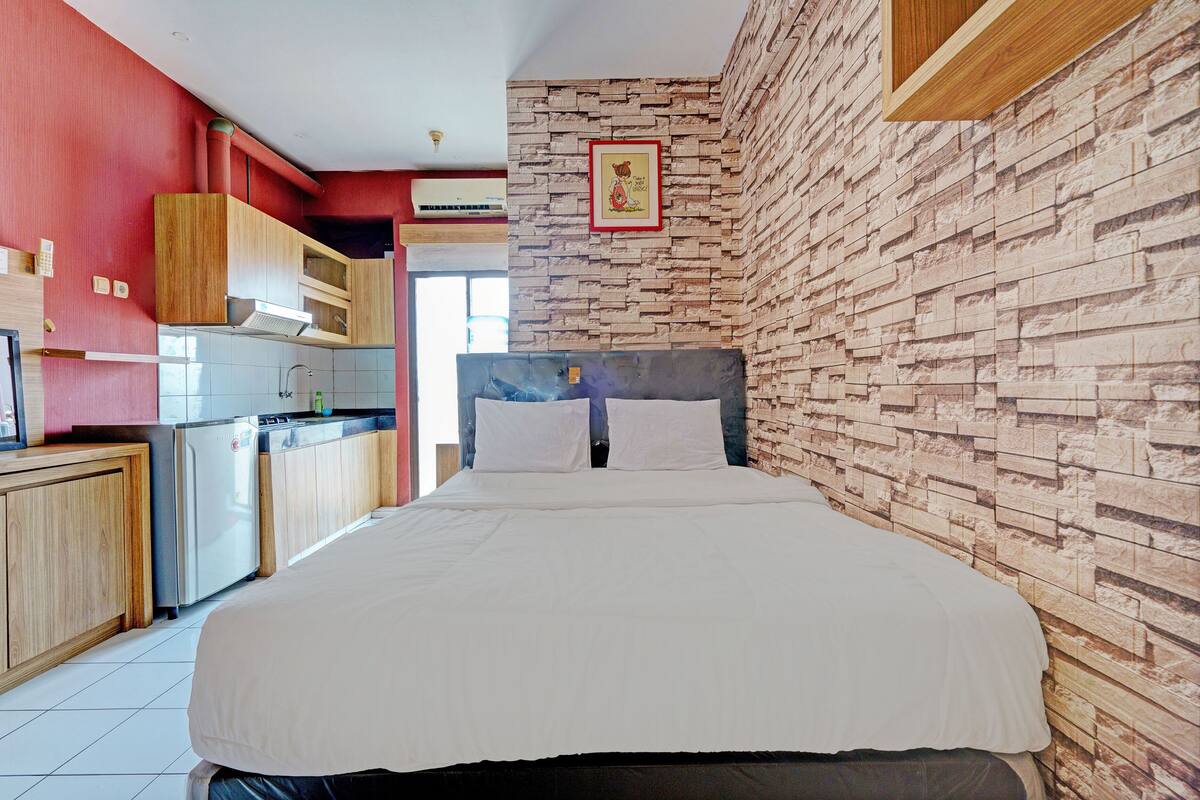 Hotelkamer met een bed, een houten hoofdbord, een muur met baksteenmotief en een kitchenette met kastjes en een koelkast.