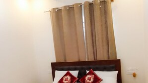 Una cama bien tendida con ropa de cama blanca y dos almohadones decorativos, un cabecero de madera, un ventilador de techo y una ventana con cortinas.