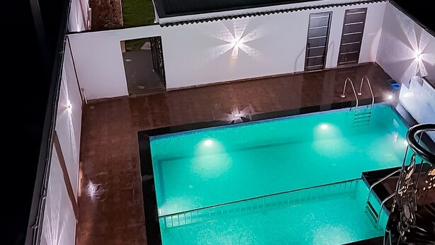 Un área de piscina moderna con una piscina rectangular, un espacio de estar cubierto y un pequeño edificio con ventanas.