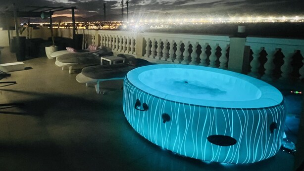 Um lounge no terraço com banheira de hidromassagem aquecida, espreguiçadeiras e vista para a cidade à noite.