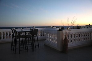 A terrace with a table and chairs, a decorative vase, and a railing overlooking a body of water during sunset.