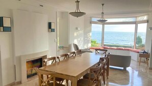 Un comedor con mesa y sillas de madera, chimenea y vistas al mar a través de amplios ventanales.