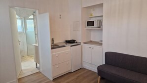 A compact kitchen with a microwave, a stove, and a sink. There is a grey sofa and a door leading to a bathroom.