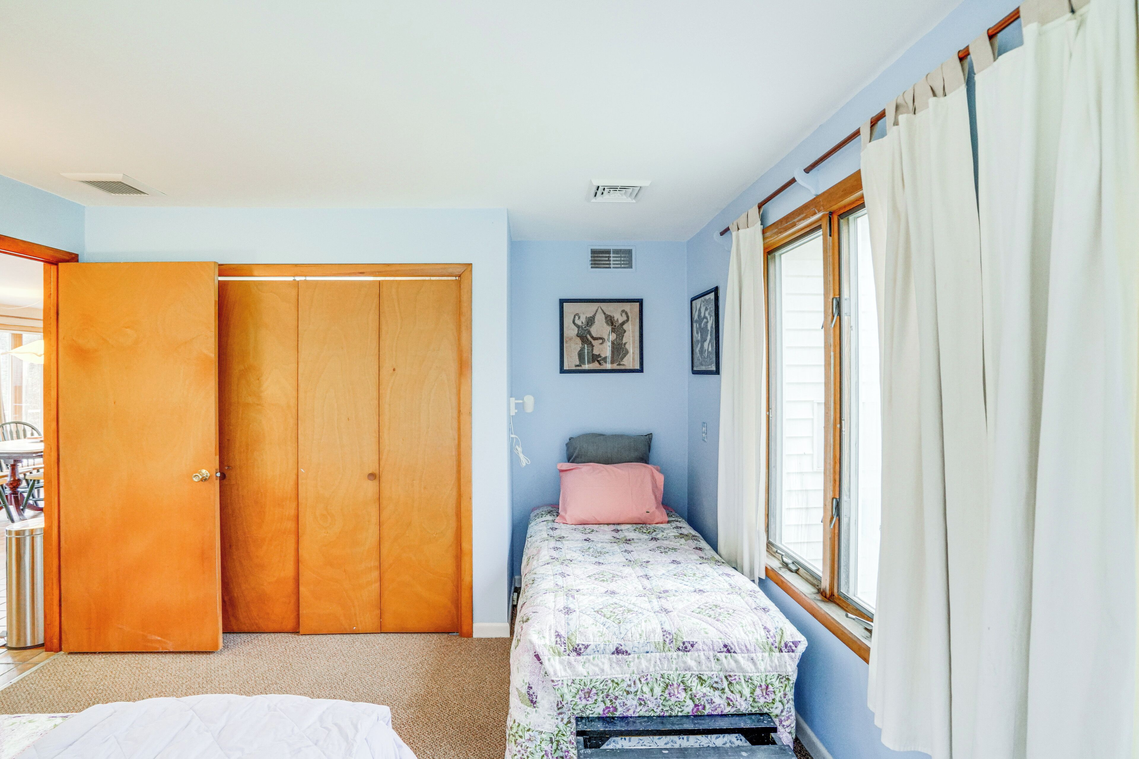 Ein Schlafzimmer mit einem Bett, einem Kleiderschrank, einem Fenster mit Vorhängen und einer Wand mit zwei gerahmten Bildern.