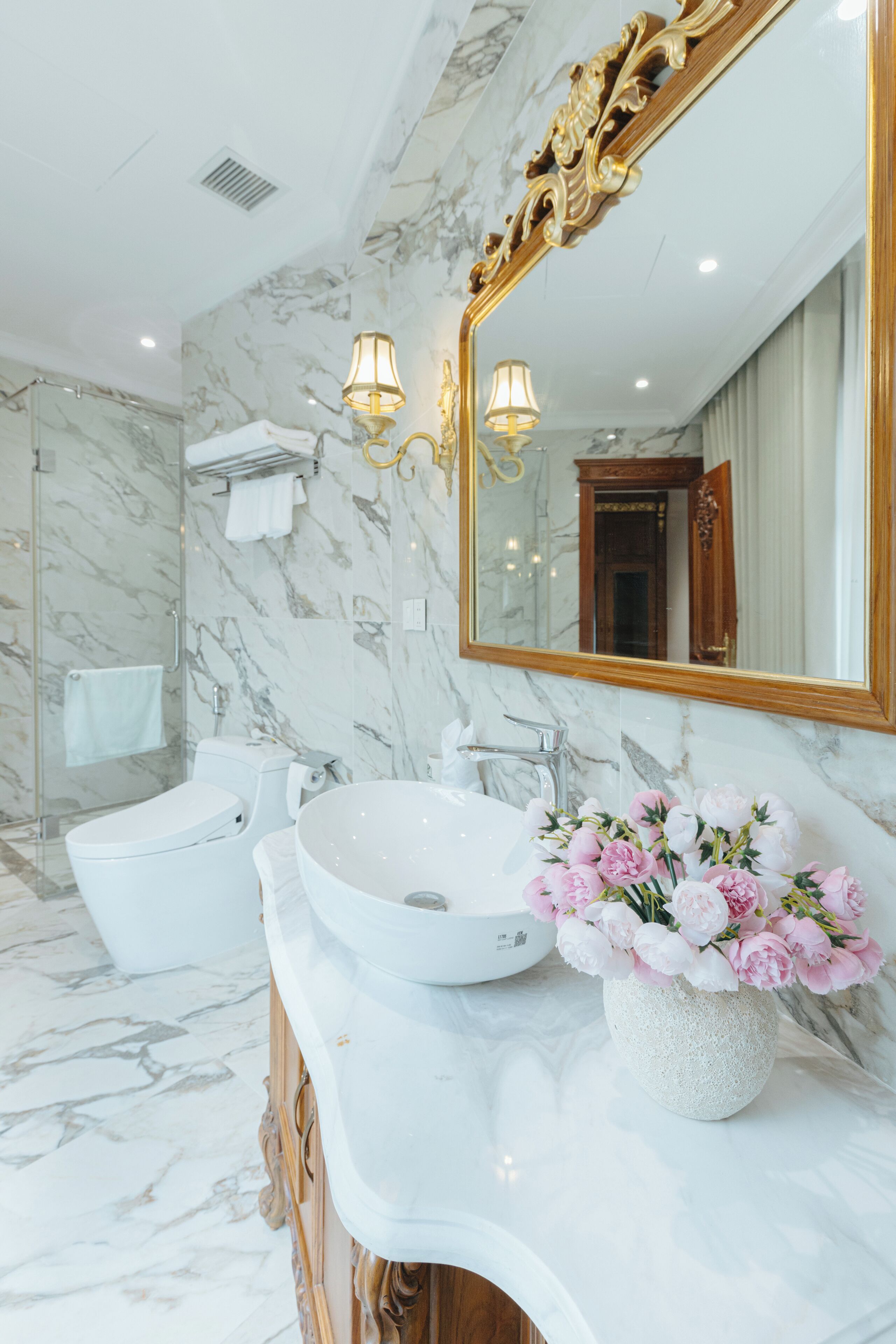 Une salle de bain avec un lavabo blanc, un grand miroir et un vase rempli de fleurs roses.