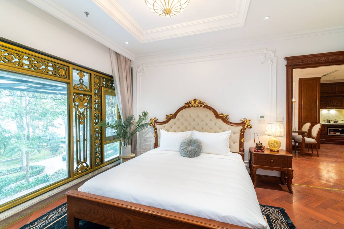 Un grand lit doté d’une tête de lit capitonnée, une table de chevet avec une lampe et une vue sur la verdure à travers une fenêtre décorative.