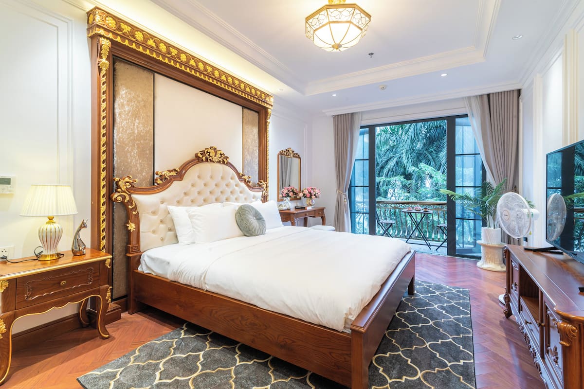 Une chambre spacieuse avec un grand lit, une table de chevet en bois, une commode, un miroir et un balcon donnant sur un paysage verdoyant.