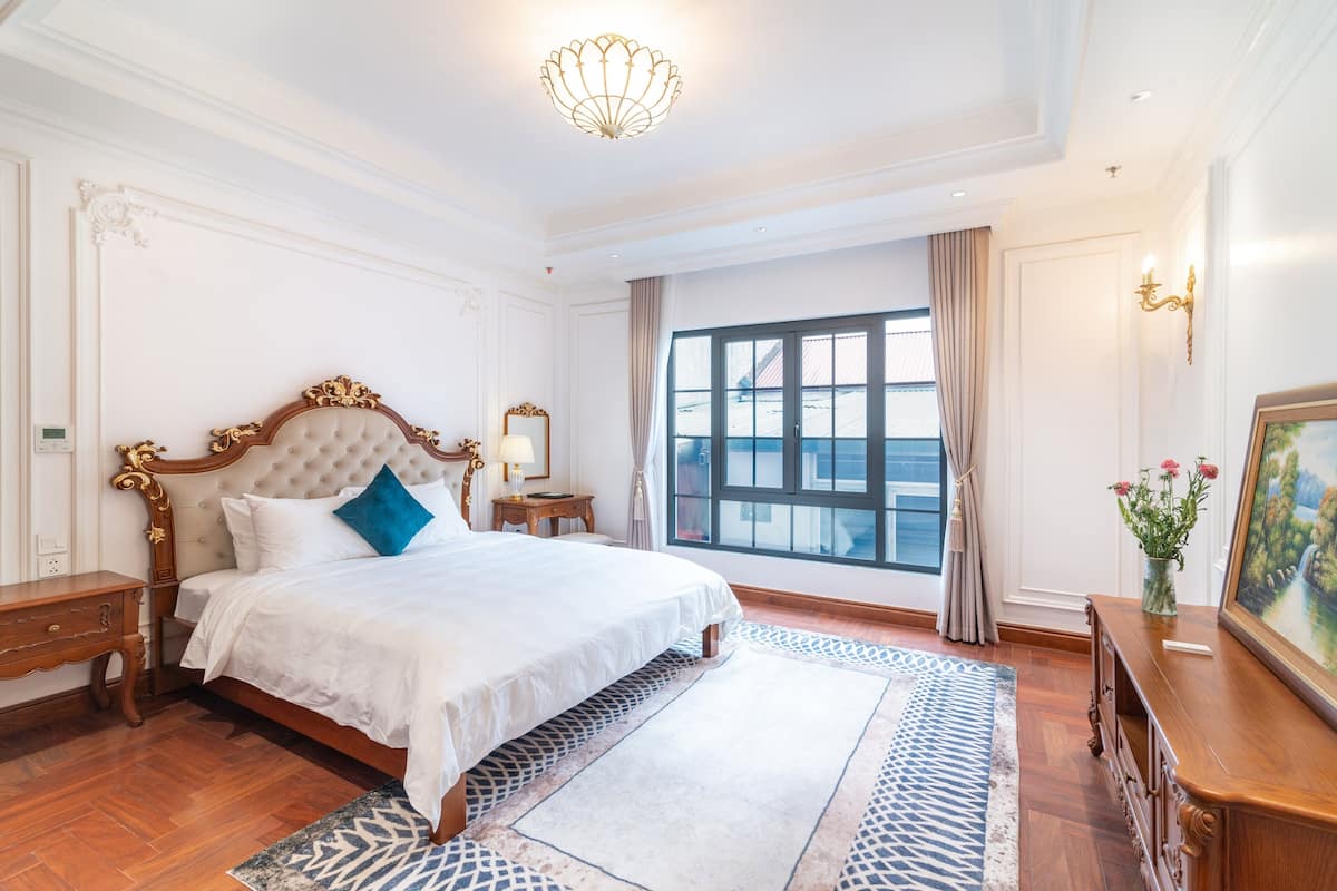 Une chambre spacieuse avec un grand lit, une table de chevet en bois, une commode, un lustre, une fenêtre avec des rideaux et un tableau accroché au mur.