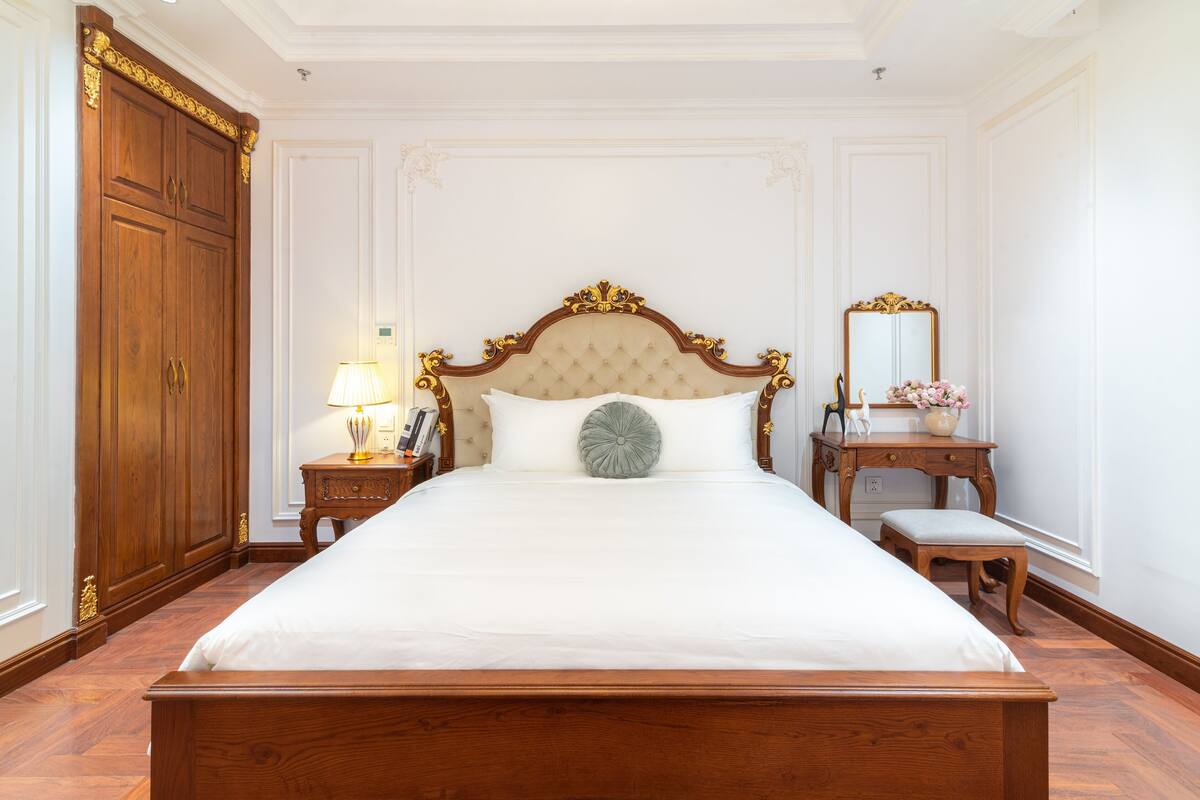 Un grand lit avec une tête de lit capitonnée, une armoire en bois, une table de chevet avec une lampe et un petit banc.
