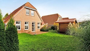 A two-story brick house with a sloped roof, a small garden, and a wooden structure to the right. - Hier Finden Sie Ruhe und Erholung in Einem Gemütlichen und Komfortablen Friesenhaus an der Nordseeküste Ostfrieslands am Weltnaturerbe Wattenmeer (Nessmersiel)