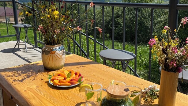 Una mesa de madera con un plato de fruta, una jarra de vidrio y un florero en un balcón con vistas a la montaña.