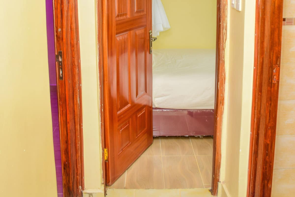 A narrow hallway with a wooden door leading to a room with a bed and a window with a white curtain.