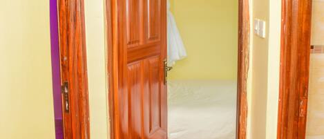 Un pasillo estrecho con una puerta de madera que da a una habitación con cama y ventana con cortina blanca.