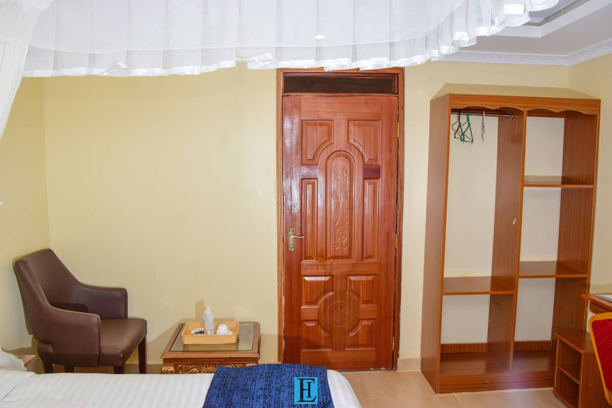 A room with a bed, a wooden wardrobe, a chair, and a small table with a basket and a tissue box.