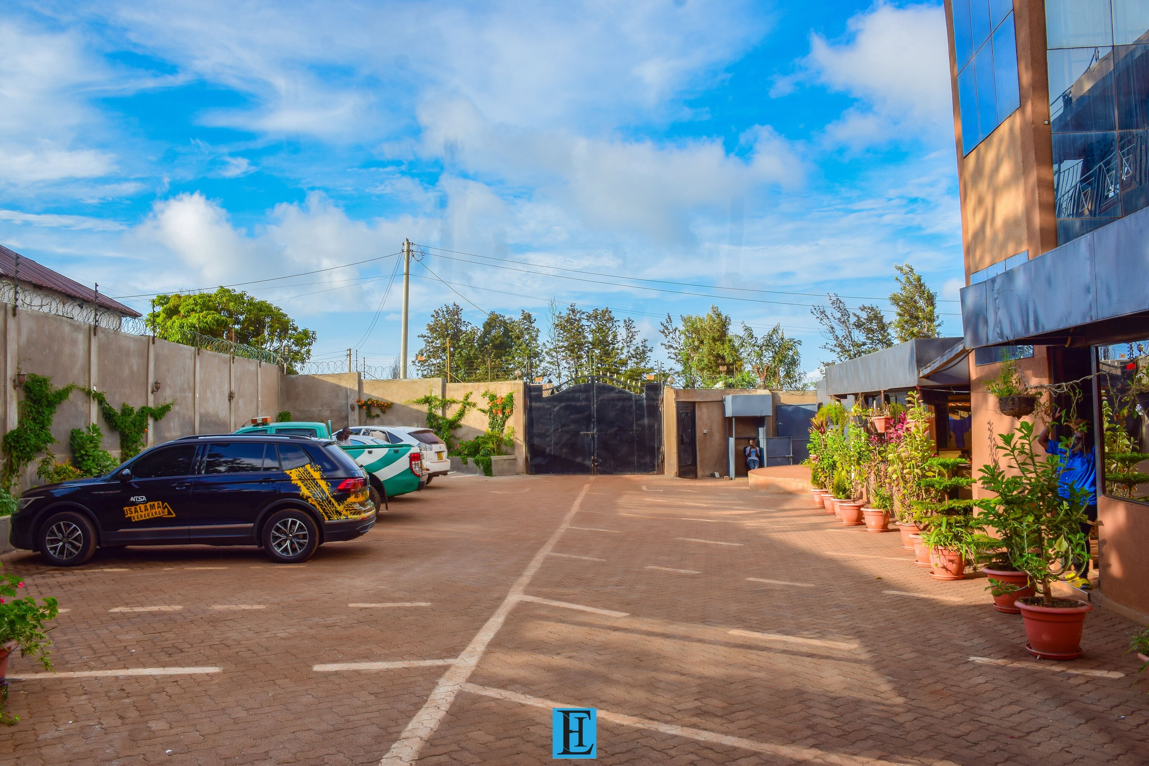 A parking area with a black SUV, a green car, and a white car parked. There are potted plants along the walkway.