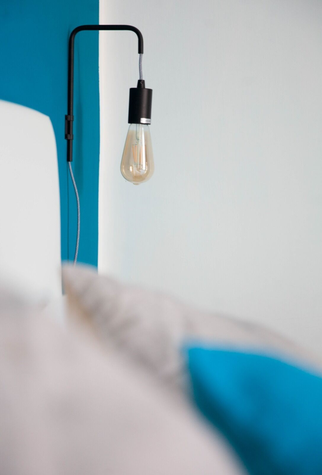 Un lit avec une tête de lit bleue adossée à un mur blanc, une ampoule suspendue et un câble noir.