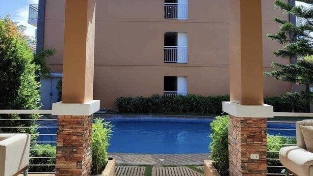 Un espacio exterior cubierto con piscina, pilares de piedra y vistas a un edificio con balcones.